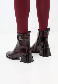 Patent leather ankle boots with a dark burgundy finish, square toe, and chunky black heel; features a decorative strap and zip closure.