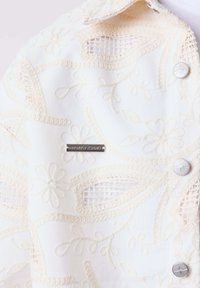 Tissu blanc avec broderie florale crème, découpes en résille, trois boutons argentés et une petite plaque métallique portant l’inscription « PROJECT X PARIS ».