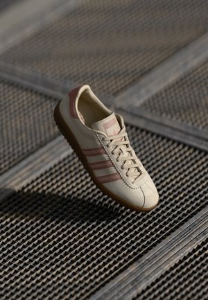 Beige Adidas sneaker met bruine strepen en rubberen zool, geplaatst op een metalen gerasterd oppervlak onder zacht licht.