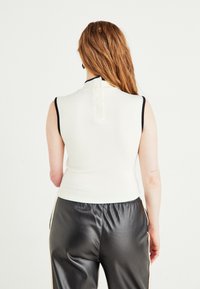 Person with long red hair wearing a white sleeveless top with black trim and black leather pants facing away against a white background.