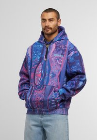 Uomo che indossa un felpa con cappuccio a fantasia multicolore blu e viola con tasche frontali e jeans azzurri chiari, in piedi contro uno sfondo semplice.