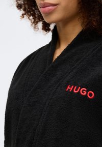 Čierny uterákový župan so šálovým golierom. Obsahuje červené vyšívané logo "HUGO" na hrudi. Mäkká textúra s prekrývajúcim dizajnom.