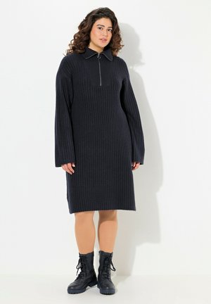 Robe côtelée bleu marine à manches longues, col à revers et fermeture éclair sur le devant. La robe tombe juste au-dessus des genoux. Associée à des bottes noires à lacets.