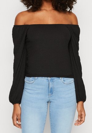 Mujer con una blusa negra de manga larga y hombros descubiertos, combinada con unos pantalones vaqueros de tiro alto en color azul claro, frente a un fondo blanco.