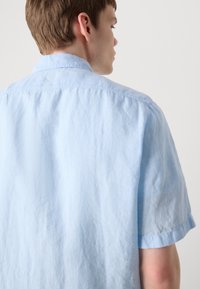 Light blue short-sleeve shirt made of textured fabric, featuring a classic collar and a relaxed fit. Visible stitching along the seams.