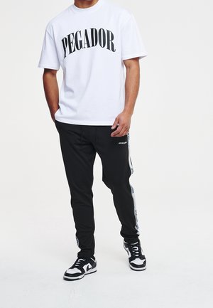 Homme portant un T-shirt blanc avec l'inscription "PEGADOR", un pantalon de sport noir à bandes latérales, et des baskets noir et blanc, debout avec une main dans la poche.