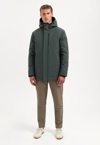 Groene geïsoleerde jas met een capuchon, voorkant met ritssluiting en drukknoopjes. Gecombineerd met beige broek en witte sneakers, minimalistisch ontwerp, zachte textuur.