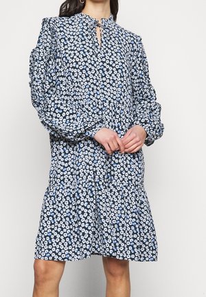 Kobieta ubrana w luźną czarną sukienkę z białym i niebieskim kwiatowym wzorem, z długimi bufkami na rękawach oraz dekoltem w kształcie klucza z wiązaniem.