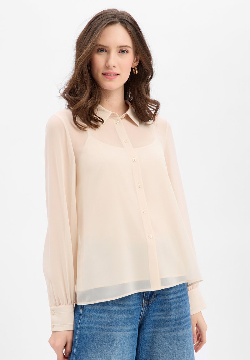 Bluzka z długim rękawem w jasnobeżowym kolorze, wykonana z przezroczystej tkaniny, z klasycznym kołnierzykiem i zapinana na guziki z przodu. Charakteryzuje się miękką teksturą i mankietami na rękawach.