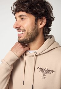 Sudadera beige con cordones, con "Pima County Tucson Arizona" bordado en marrón. Tejido suave, puños de canalé y corte relajado.