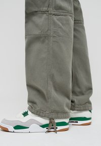 Zapatillas deportivas blancas y verdes con acentos grises, combinadas con pantalones holgados color verde oliva que tienen costuras visibles y un ajuste relajado.