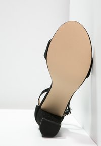 Sandale à talons en daim noir avec une bride à la cheville, montrant la texture lisse de la semelle beige en caoutchouc et la surface texturée pour l'adhérence.