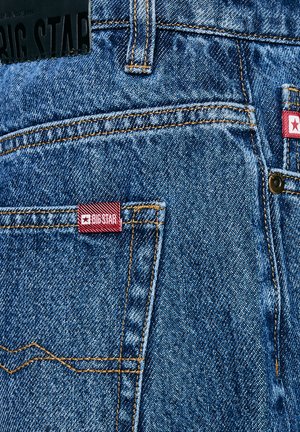 Kék farmernadrág, bőr márkajelzéssel a deréknál, piros szövetcímkével, rajta logóval, és kontrasztos sárga cérnával a zsebeknél.