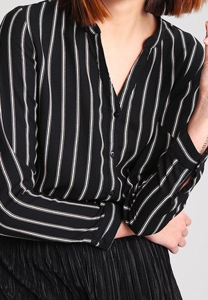 Osoba ubrana w czarną koszulę z długim rękawem z białymi pionowymi paskami oraz czarne plisowane spodnie, z założonymi skrzyżowanymi rękami, zdjęcie przycięte na wysokości ramion.