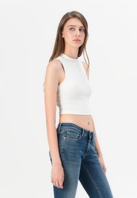 Jauna sieviete, tērpusies baltā bezpiedurkņu topā un zilās džinsās, stāv labajā pusē pret vienkāršu baltu fona, skatoties sāņus.