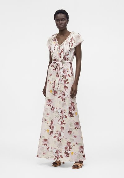 Γυναίκα που φοράει αμάνικο, μακρύ φόρεμα με λουλούδια, με βολάν στο V-λαιμό και ζώνη στη μέση, σε συνδυασμό με μπεζ σανδάλια με τακούνι, στέκεται μπροστά σε λευκό φόντο.