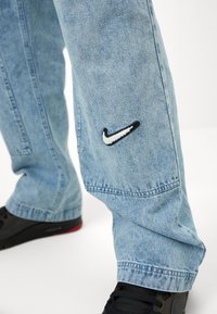 Világoskék farmernadrág fekete-fehér hímzett Nike logóval a bal lábon, laza szabású, látható varrási részletekkel.