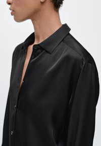 Black satin shirt featuring a classic collar, front button closure, and smooth texture, with a relaxed fit and gently curved shoulder seams.
