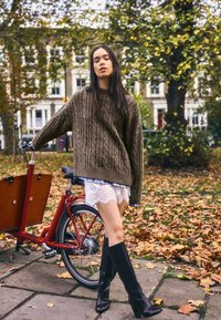 Barna kötött pulóver, fehér csipkés rövidnadrág, fekete térdig érő csizma. Egy piros teherbicikli mellett áll, körülötte őszi levelek.