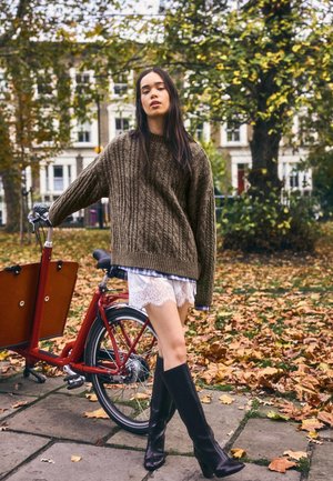 Maglione marrone a trecce, pantaloncini bianchi in pizzo, stivali neri fino al ginocchio. In piedi accanto a una bicicletta cargo rossa, circondata da foglie autunnali.