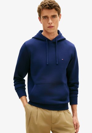 Young man wearing a navy blue hoodie with front pocket and beige pants, standing with one hand in pocket against a plain background.