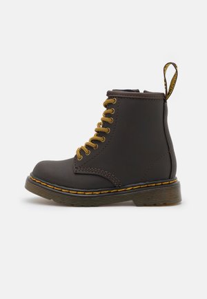 Donkerbruine leren enkellaars met gele veters, gele stiksels langs de zool en een merkpull-tab aan de achterkant op een witte achtergrond.