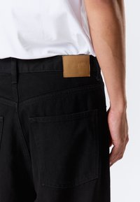 Pantalon en denim noir avec une coupe droite, doté de deux poches arrière et d'un patch en cuir tan sur la taille avec un texte en relief.