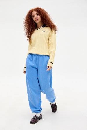 Jeune femme aux cheveux roux bouclés portant un pull jaune avec un col marine, un pantalon bleu ample, des chaussettes blanches et des mocassins marron foncé marchant en avant.