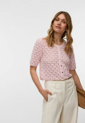Femme aux cheveux blonds ondulés portant un cardigan court en tricot rose clair boutonné et un pantalon crème taille haute, tenant un sac marron.