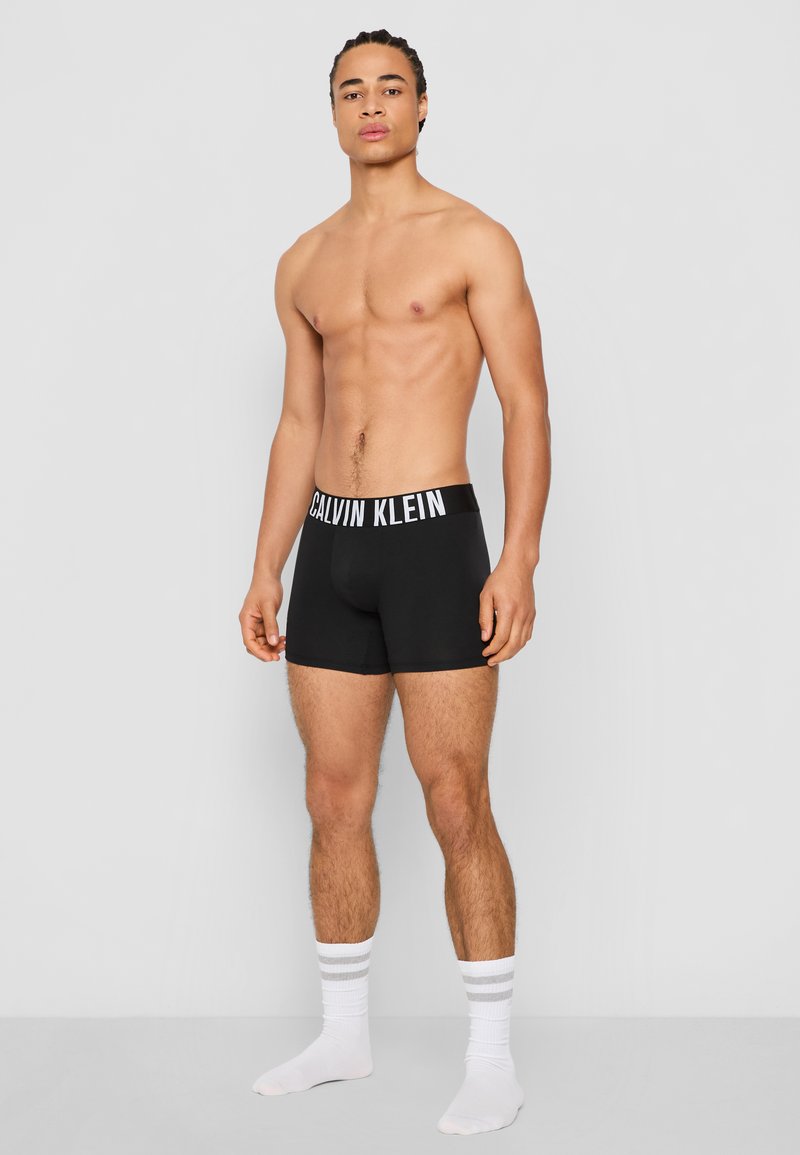 Crne bokserice s bijelim pojasom s Calvin Klein logom. Model stoji bos, noseći bijele čarape sa sivim prugama. Glatka tekstura tkanine.