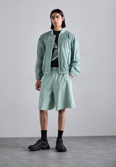 Veste légère vert menthe avec des poignets élastiques, shorts assortis en vert menthe et baskets noires à semelles épaisses. Le motif principal sur le t-shirt est sombre.