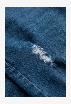 Blauwe denimstof met een versleten stukje dat rafelige randen vertoont. De textuur is glad, met zichtbare weefpatronen in het materiaal.