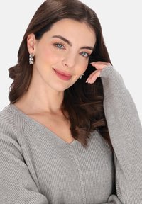 Szürke kötött pulóver V-nyakkal, bordázott textúrával és hosszított ujjakkal. A modellen ezüst karika fülbevalók is láthatók.