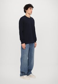 Sweatshirt azul-marinho com decote redondo e punhos canelados, combinado com jeans de perna larga azul-claro e sapatilhas brancas, posando frente a um fundo simples.
