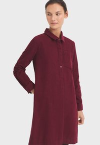 Vestido de camisa color burdeos fabricado en un tejido suave, con cuello, parte delantera abotonada y mangas largas, y un bolsillo en la parte izquierda.