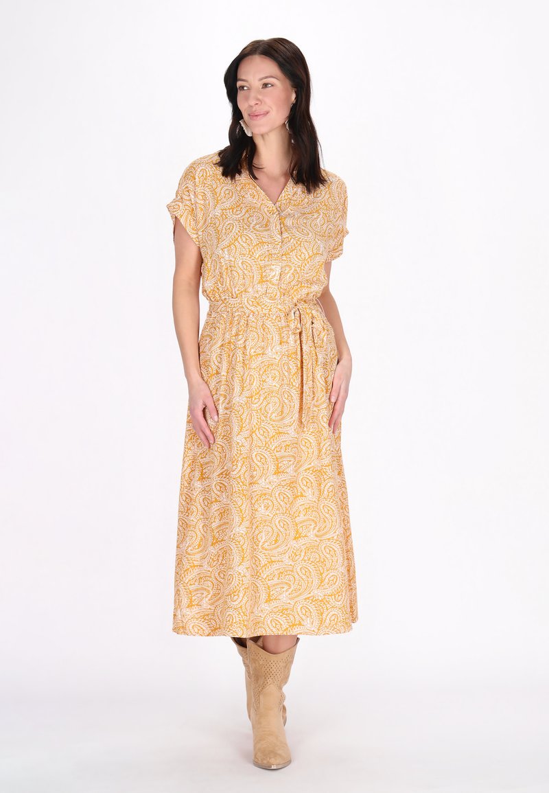 Abito giallo a fantasia paisley con maniche corte, vita sfiancata e cintura con fiocco. Abbinato a stivali da cowboy color beige chiaro.
