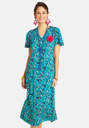 Woman in teal floral dress with short sleeves, pink flower pin, large pink earrings, blue beaded necklace, and gold bracelets smiling.