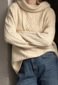 Maglione a collo alto di colore crema con lavorazione a trecce, caratterizzato da un motivo a diamante. Indossato sopra jeans in denim blu. Texture morbida.