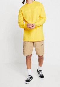 Maglietta gialla a maniche lunghe, pantaloni cargo beige, scarpe da ginnastica nere con suole bianche e calze. Design semplice con una stampa sul torso e tasca laterale.