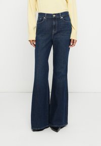 Jean en denim bleu foncé évasé avec poches avant, fermeture par boutons et passants pour ceinture ; associé à un haut jaune clair.