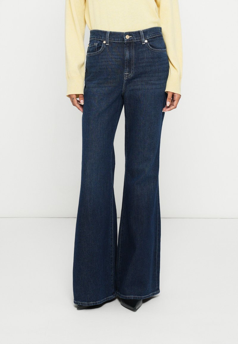 Jean en denim bleu foncé évasé avec poches avant, fermeture par boutons et passants pour ceinture ; associé à un haut jaune clair.