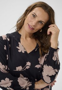 Crna cvjetna bluzica s ružičastim uzorcima, V-izrezom i nabranim manžetama. Izrađena od mekanog materijala, ima duge rukave slobodnog kroja.