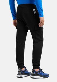 Schwarze Cargo-Jogginghosen mit Seitentaschen, elastischem Bund und reflektierenden Streifen. Getragen mit schwarzen und blauen Sportschuhen.
