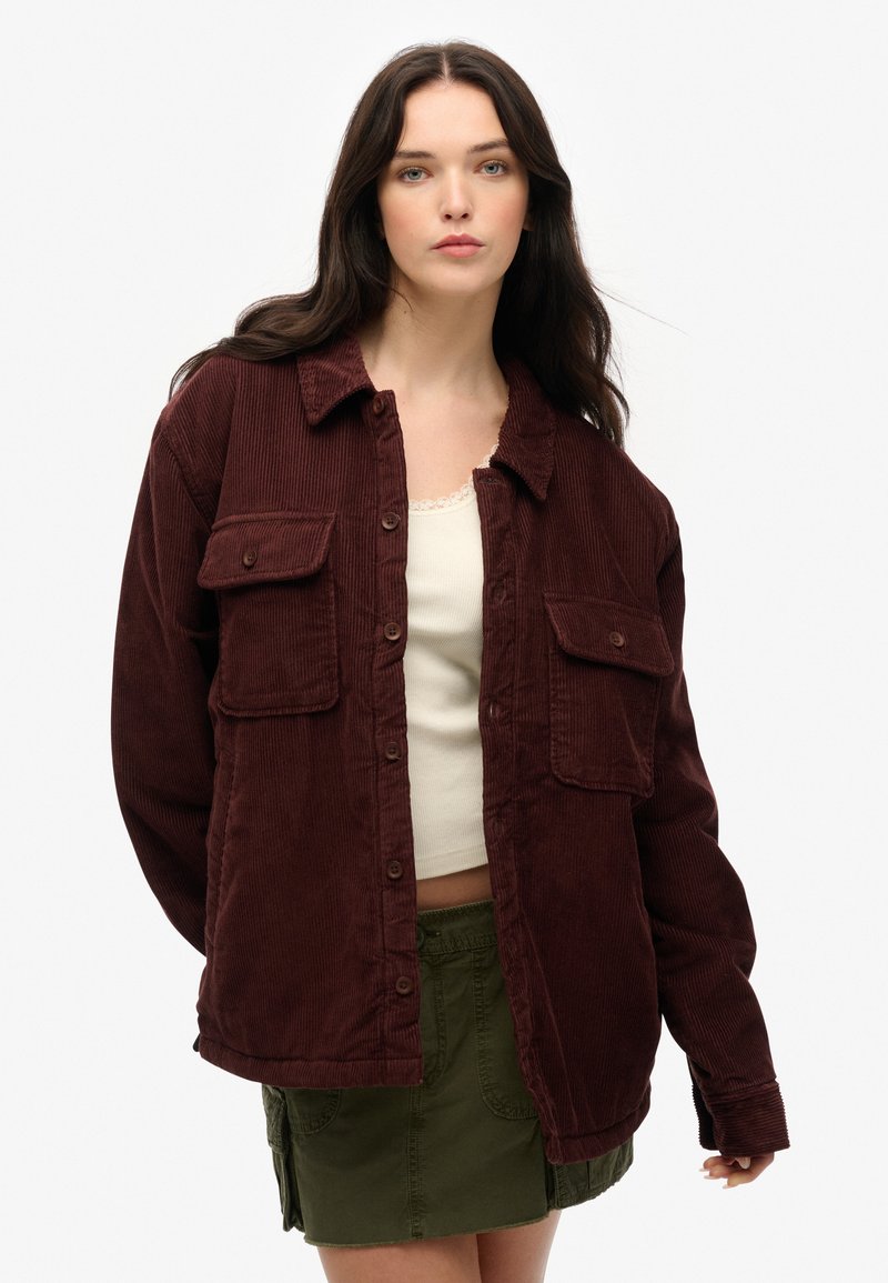 Bordeaux corduroy shirtjack met twee voorzakken, knoopsluiting en een relaxte pasvorm, gecombineerd met een witte tanktop en groene rok.