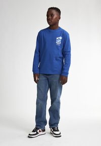 Langärmliges blaues Baumwollhemd mit weißem Druck, kombiniert mit blauen Jeans und schwarz/roten Turnschuhen. Es hat einen lockeren Schnitt und einen runden Halsauschnitt.