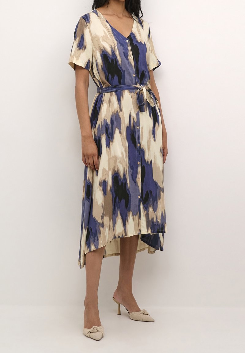 Femme portant une robe midi à manches courtes boutonnée avec un imprimé abstrait bleu, beige et noir, ceinture nouée et mules beige à talons hauts.