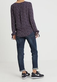 Donna con una camicetta lunga a maniche viola a motivo, jeans scuri arrotolati e sneakers nere Nike, in piedi con la schiena rivolta in avanti.