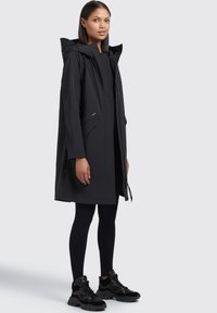 Parka nera impermeabile con cappuccio, tasche frontali e chiusura con zip. Indossato sopra leggings neri e sneakers nere. Texture liscia.