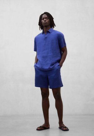 Mężczyzna o ciemnej karnacji i dredach, ubrany w niebieską koszulę z krótkim rękawem i kołnierzykiem oraz dopasowane szorty, stojący z rękami w kieszeniach i w klapkach.