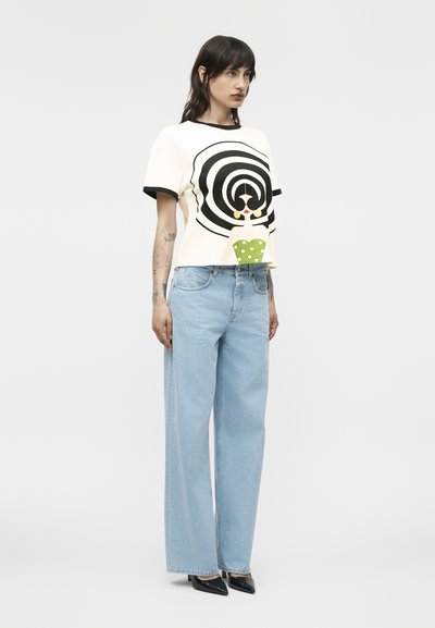 Tânără femeie stând, purtând un top alb cu mâneci scurte, cu un design negru în spirală și o figură verde cu buline, blugi evazați bleu deschis și pantofi cu toc negri.